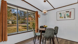 A dining area with a table and chairs, a window with curtains, a clock, and a framed abstract painting on the wall.