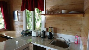 A kitchen with wooden walls, a window, a sink, a toaster, a kettle, and a shelf with glassware. - Red Hibiscus Villa 1 (Avarua)