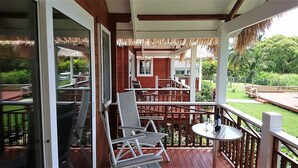 A covered outdoor area with a wooden deck, white chairs, a table with a bottle and glass, and a view of a lush garden and palm trees. - Red Hibiscus Villa 1 (Avarua)
