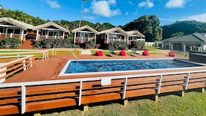 A pool area with wooden deck seating, thatched-roof structures, and a clear blue sky. - Red Hibiscus Villa 1 (Avarua)