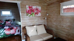 A cozy room with a wicker sofa, a bed with floral bedding, and a wall-mounted painting of pink flowers. - Red Hibiscus Villa 1 (Avarua)