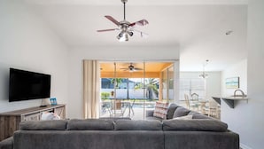 Un salon avec un canapé gris, un téléviseur à écran plat, un ventilateur de plafond et une vue sur une terrasse extérieure.