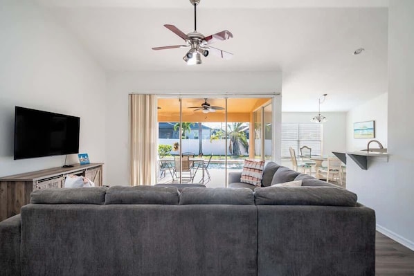A living room with a grey sofa, a flat-screen TV, a ceiling fan, and a view of an outdoor patio.
