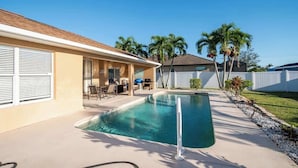 Un espace piscine résidentiel comprenant une maison, du mobilier d’extérieur et des palmiers.