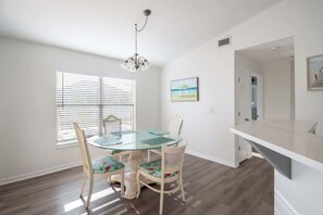 A dining area with a glass table and chairs, a chandelier, a window with blinds, and a painting on the wall.