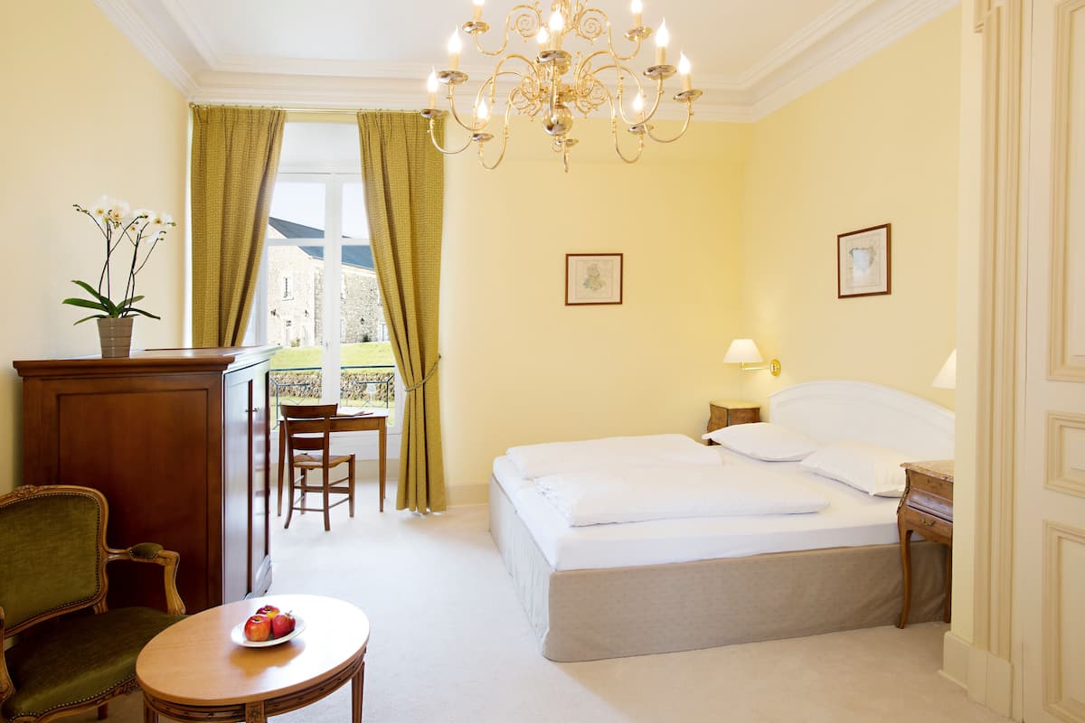 A hotel room with a bed, a desk with a chair, a dining table with a fruit plate, a chandelier, and two framed pictures on the wall.