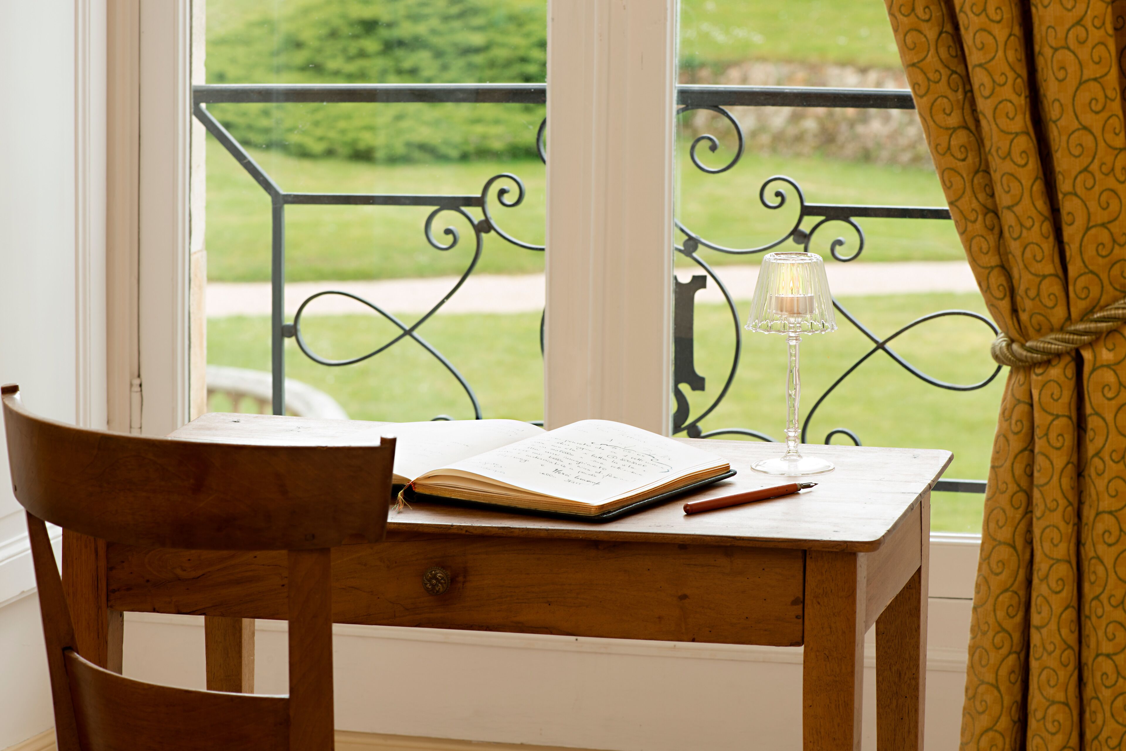 A wooden desk with an open book and a glass of water, positioned by a window with a view of a green landscape.