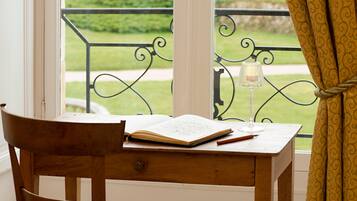 A wooden desk with an open book and a glass of water, positioned by a window with a view of a green landscape.
