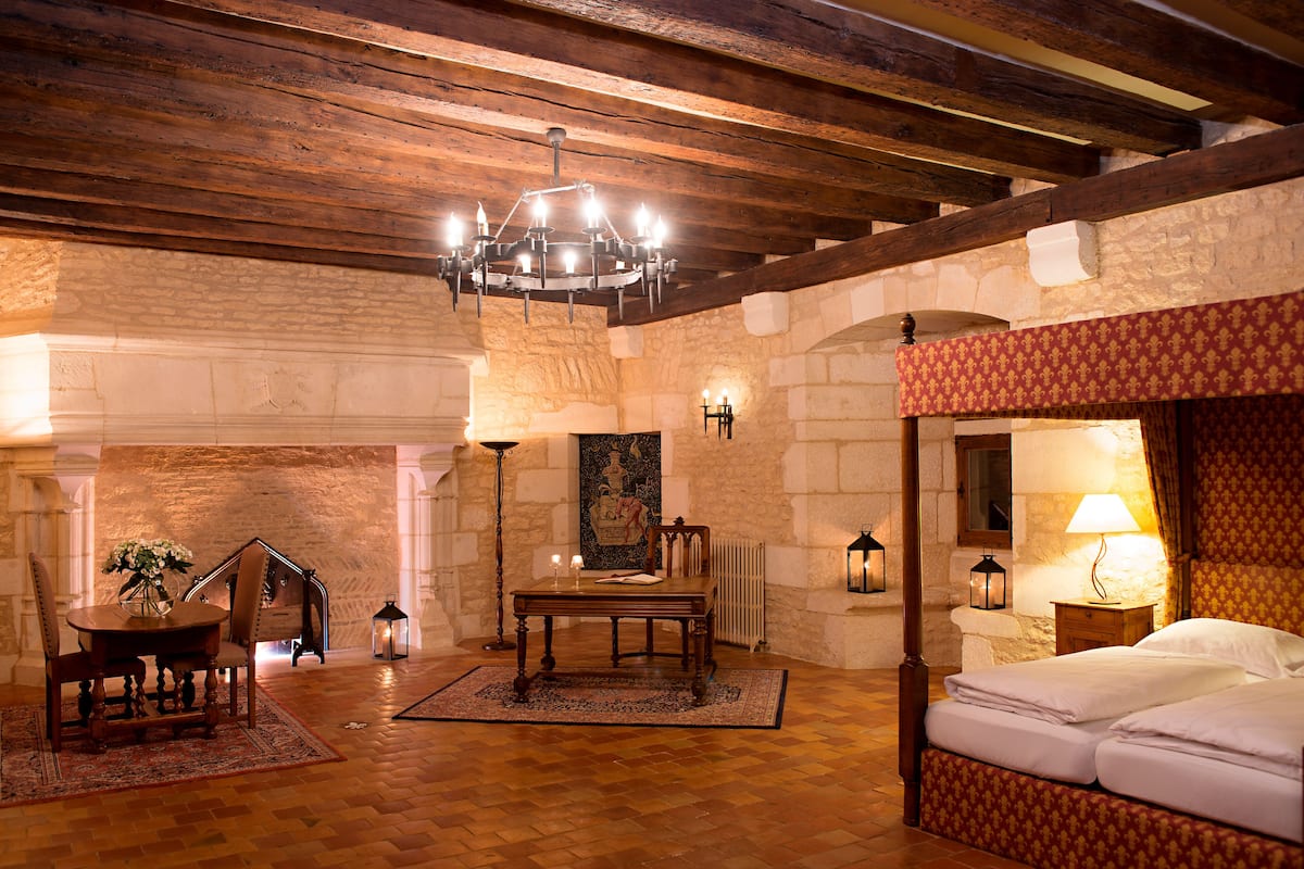 A room with a wooden ceiling, stone walls, a bed with a red headboard, a table with chairs, and a chandelier.