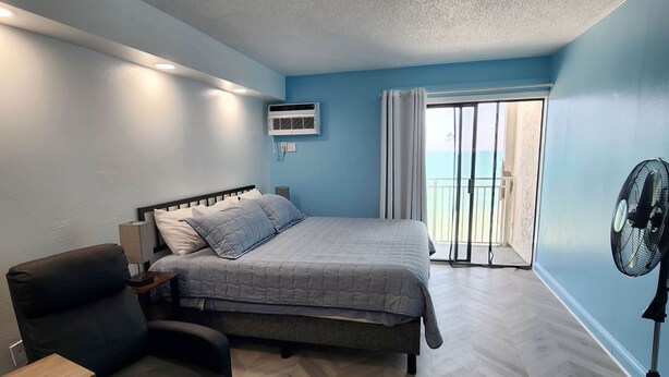 A bedroom with a bed, a chair, a desk, an air conditioner, and a fan. - Oceanfront Efficiency T604 (Myrtle Beach)