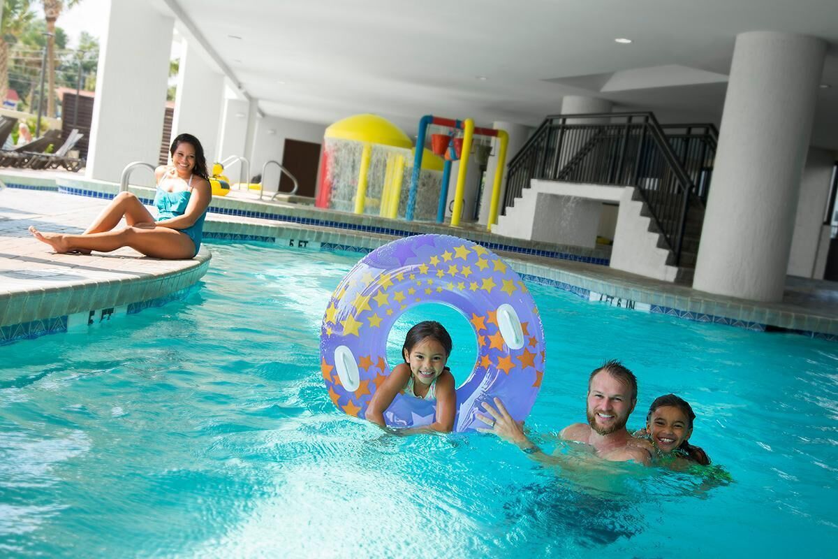 Ein Schwimmbecken mit Kind und Erwachsenem im Wasser, ein Liegebereich mit Sonnenliegen und ein überdachter Bereich mit Treppe und Geländer.