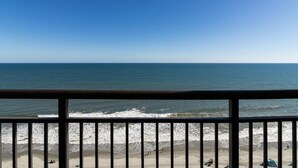 Vista de la playa desde un balcón con barandilla, olas del océano y cielo despejado.