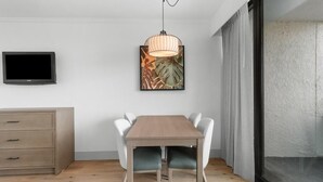 Uma área de refeições moderna com mesa de madeira e cadeiras brancas, uma televisão montada na parede e uma obra de arte emoldurada acima da mesa.