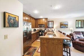 A kitchen with wooden cabinets, a stainless steel refrigerator, a microwave, and a stove. There is a dining area with a brown leather couch and a table. - Evergreens 1221- Mtn Condo, Gated Resort, Indoor/Outdoor Pools & Hot Tub (Boone)