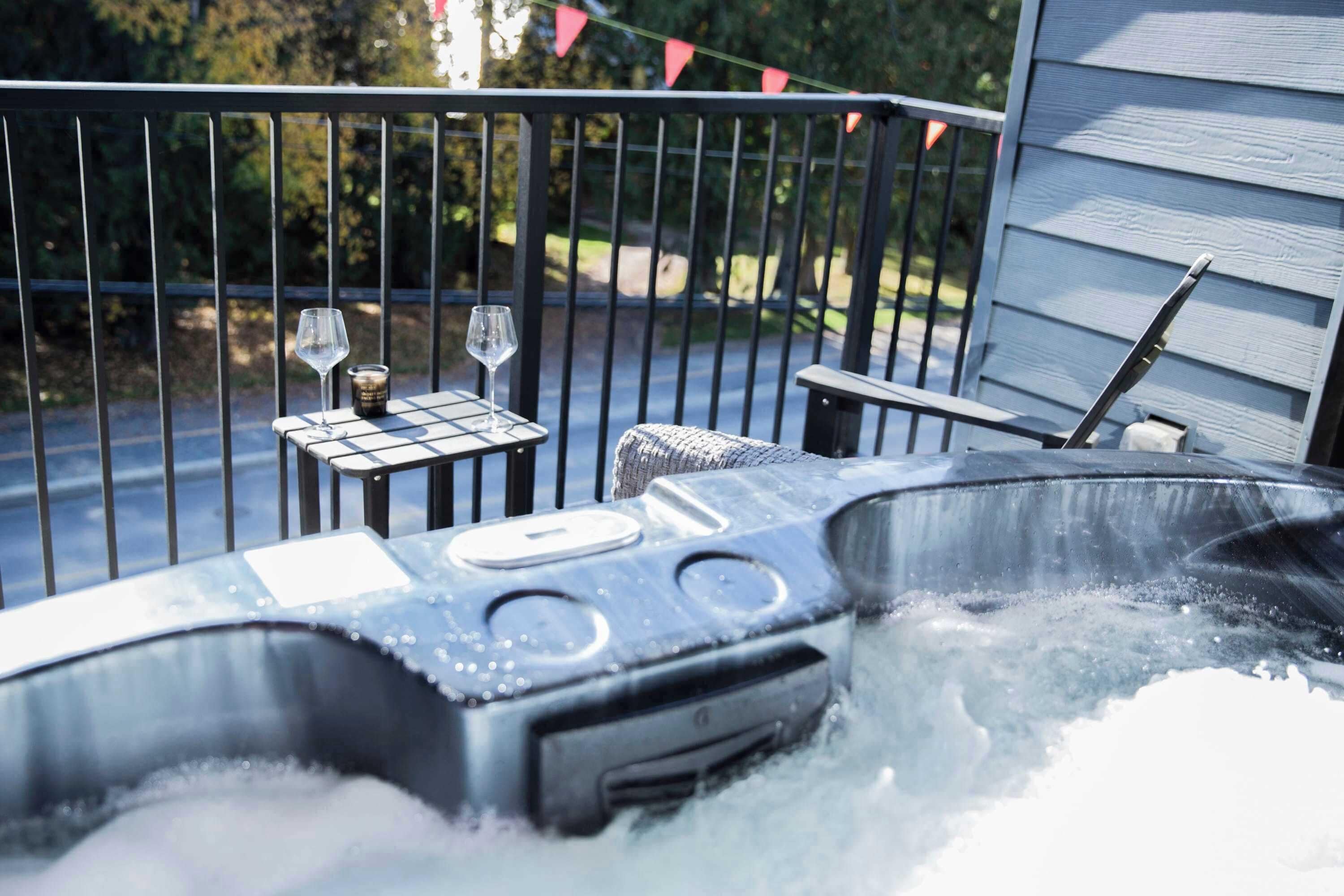 A hot tub with two glasses and a container on a table, surrounded by a metal fence and a house in the background.