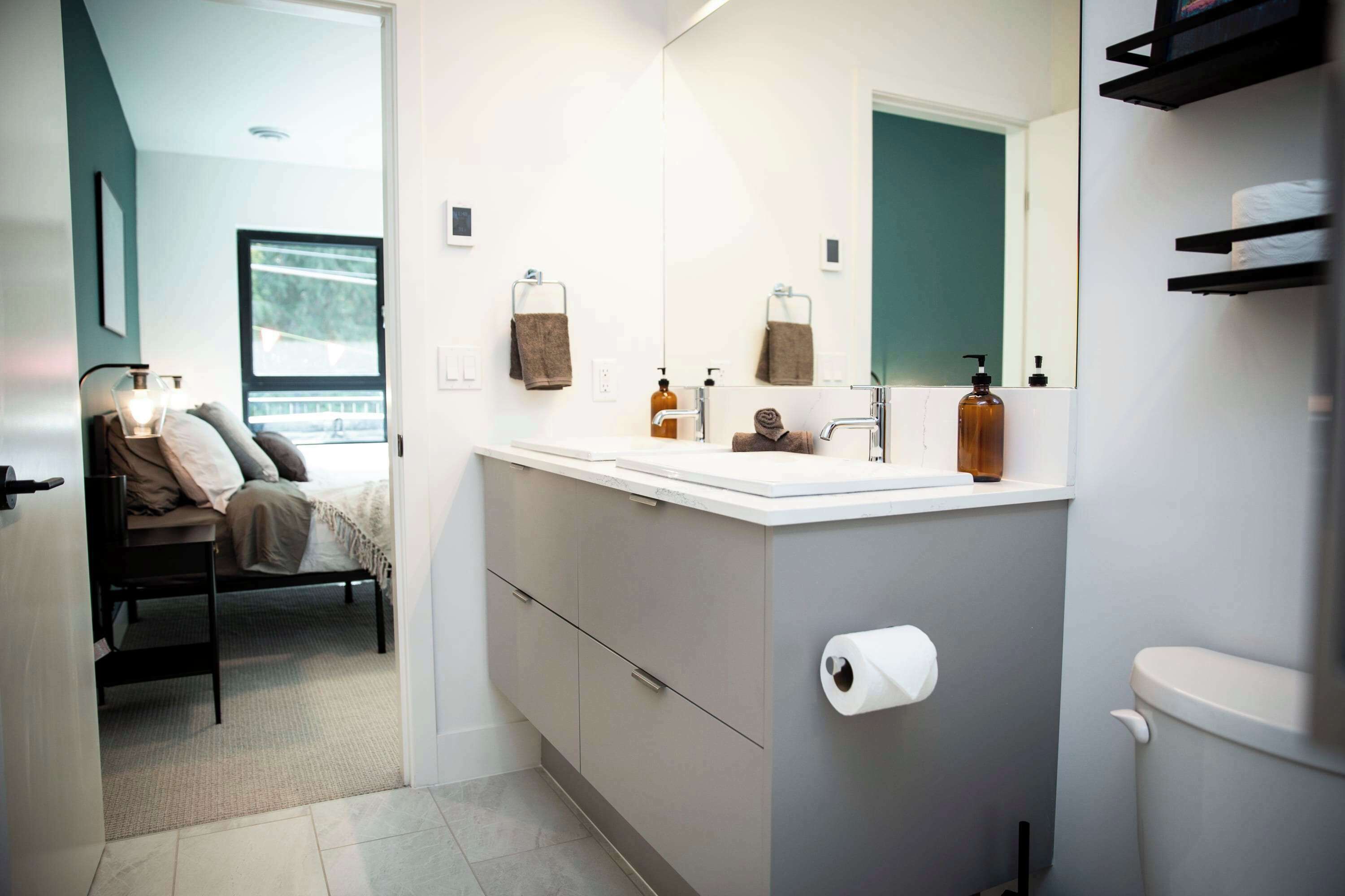 A modern bathroom with a large mirror, a toilet, and a sink. There is a bed with a view of the outdoors visible through a window.