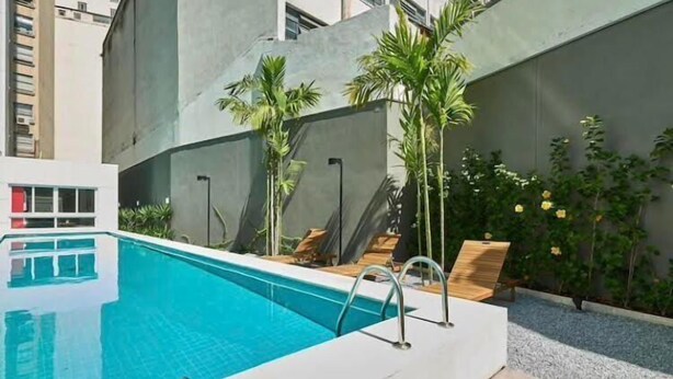 A modern outdoor pool area with a clear blue pool, surrounded by palm trees and a concrete wall. - Luz 1802 (São Paulo)