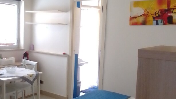 A hotel room with a bed, a desk, and a chair. There is a window with blinds, a door, and a painting on the wall.