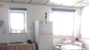 A small, well-lit room with a kitchenette, a refrigerator, a table with chairs, and a bed with a blue cover.
