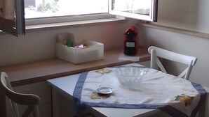 A small dining area with a table set for two, a window with blinds, a red kettle, and a white chair.