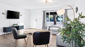 Ein modernes Wohnzimmer mit einem Flachbildfernseher, einem dunkelgrauen Sofa, zwei schwarzen Sesseln, einem kleinen runden Tisch und einer großen Stehlampe.