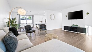 Ein modernes Wohnzimmer mit einer Couch, Sesseln, einem Couchtisch, einem Fernseher und einem Essbereich im Hintergrund.