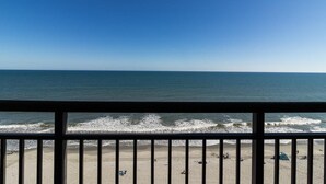 Une vue sur la plage depuis un balcon doté d’une balustrade, avec les vagues de l’océan et un ciel dégagé.