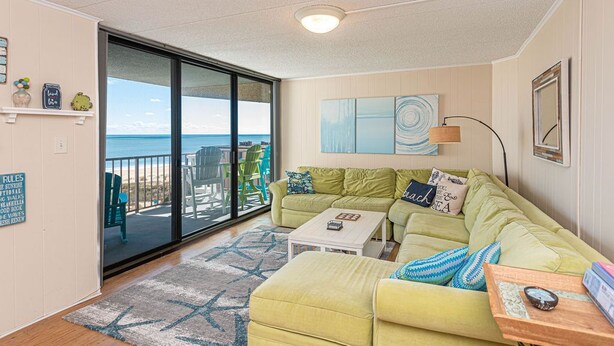 A living room with a green sofa, a coffee table, and a view of the beach through large sliding glass doors.