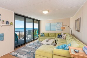 A living room with a green sofa, a coffee table, and a view of the beach through large sliding glass doors. - Spacious, comfy 2 bedroom south side oceanfront condo with free WiFi, an outdoor pool, and a pretty ocean view located uptown just steps to the beach! (Ocean City)