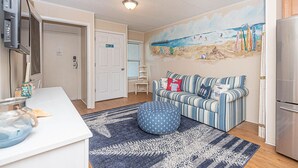 A cozy living room with a striped sofa, a blue ottoman, and a beach-themed mural on the wall.