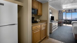 A compact kitchen with wooden cabinets, a microwave, and a coffee maker. A dining area with a table and chairs is visible, leading to a balcony with a view. - Unforgettable Moments 7th Floor Two Queen Room (Myrtle Beach)