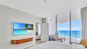 Una habitación de hotel moderna con un televisor grande mostrando una playa tropical, un balcón con vista al océano y un diseño minimalista.
