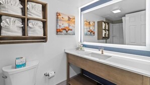 A bathroom with a white toilet, a sink with a mirror, and a shelf with folded towels. - Low Flr Oceanfront Deluxe Queen Parlor Efficiency (Myrtle Beach)