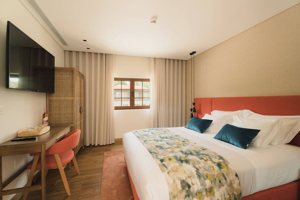 A hotel room with a bed, a desk with a chair, a television, and a window with curtains.