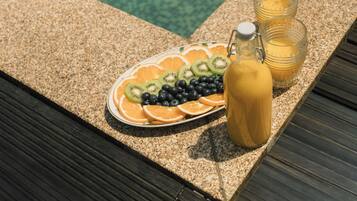 A poolside setting with a fruit platter, two glasses of juice, and a bottle of juice on a wooden deck.