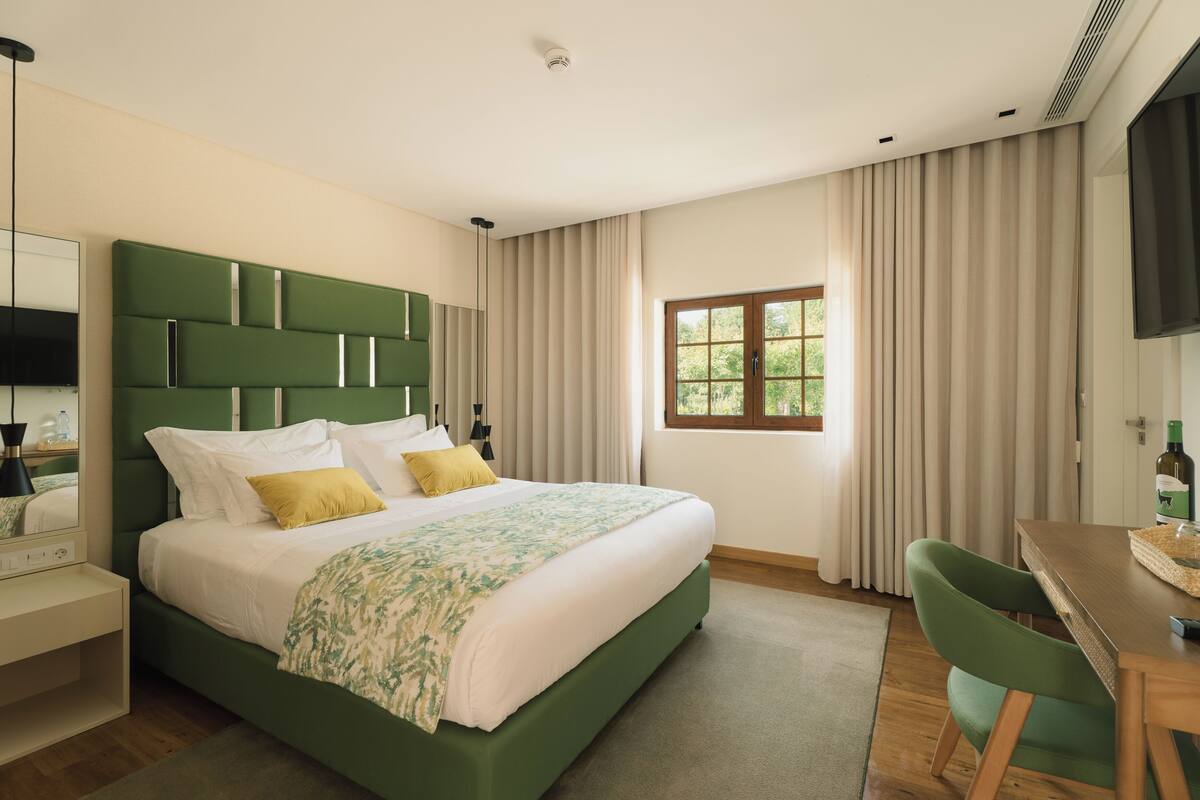 A modern bedroom with a green bed, white bedding, a desk, and a window with curtains.
