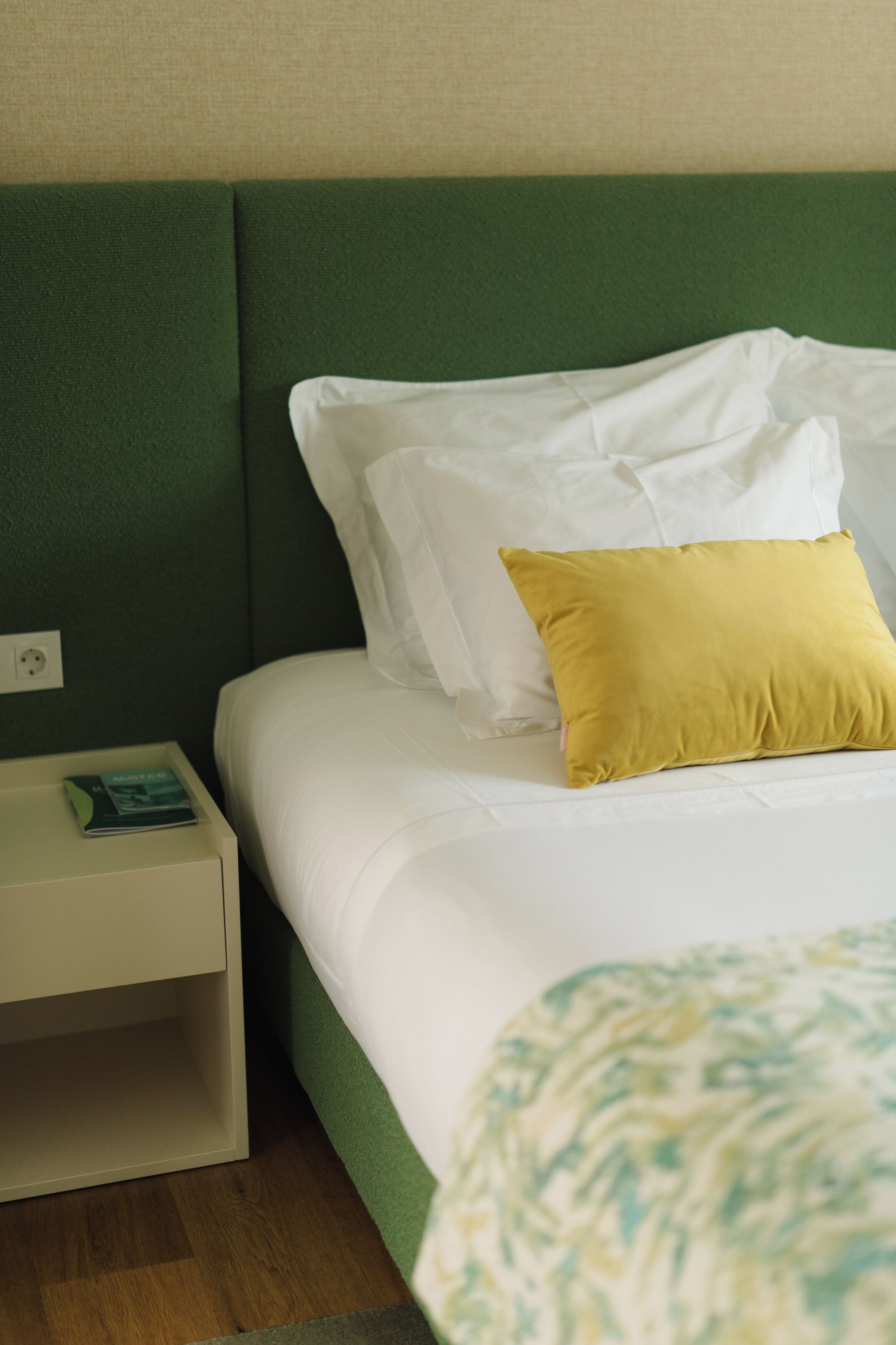 A neatly made bed with white and yellow pillows, a green headboard, and a bedside table with a book.