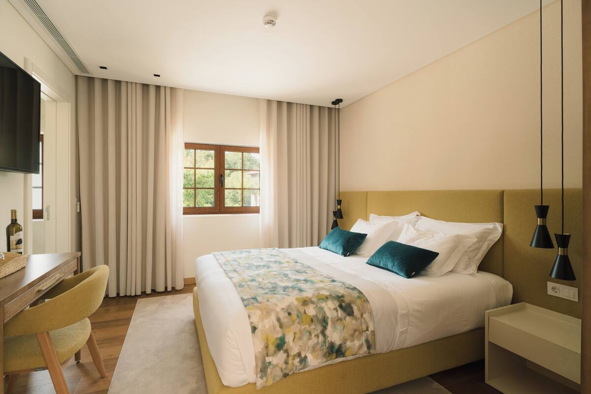 A bedroom with a bed, a desk, a chair, a window, and a TV.
