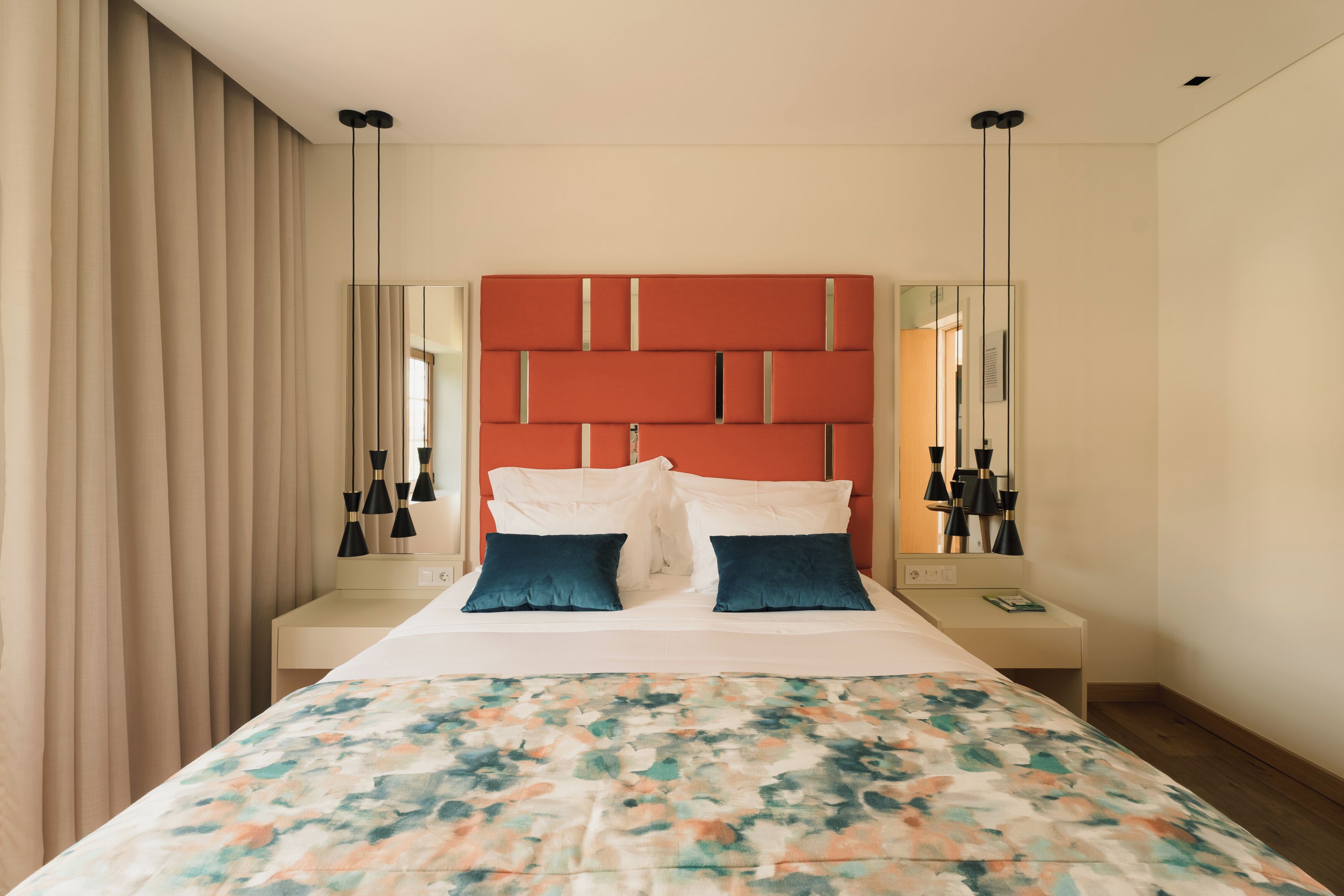 A hotel room with a bed, a bedside table, a nightstand, a mirror, and a window with curtains.