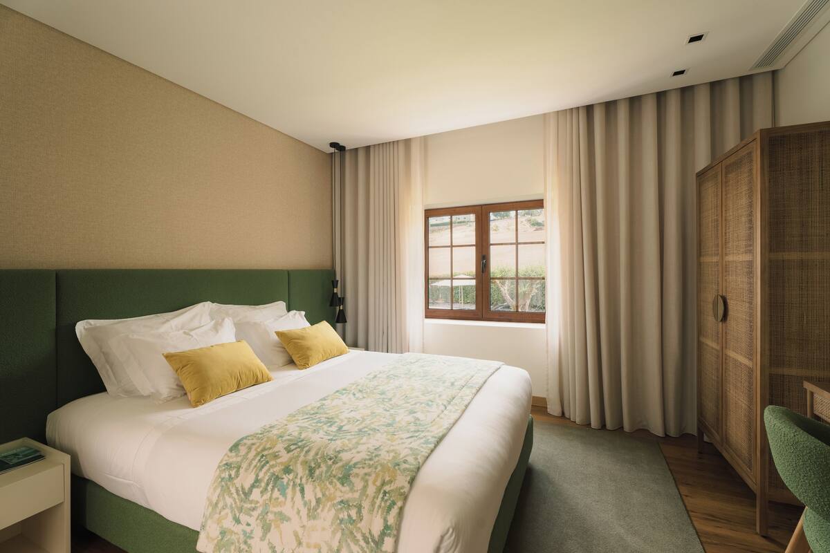 A hotel room with a large bed, a nightstand, a green chair, a wardrobe, and a window with curtains.