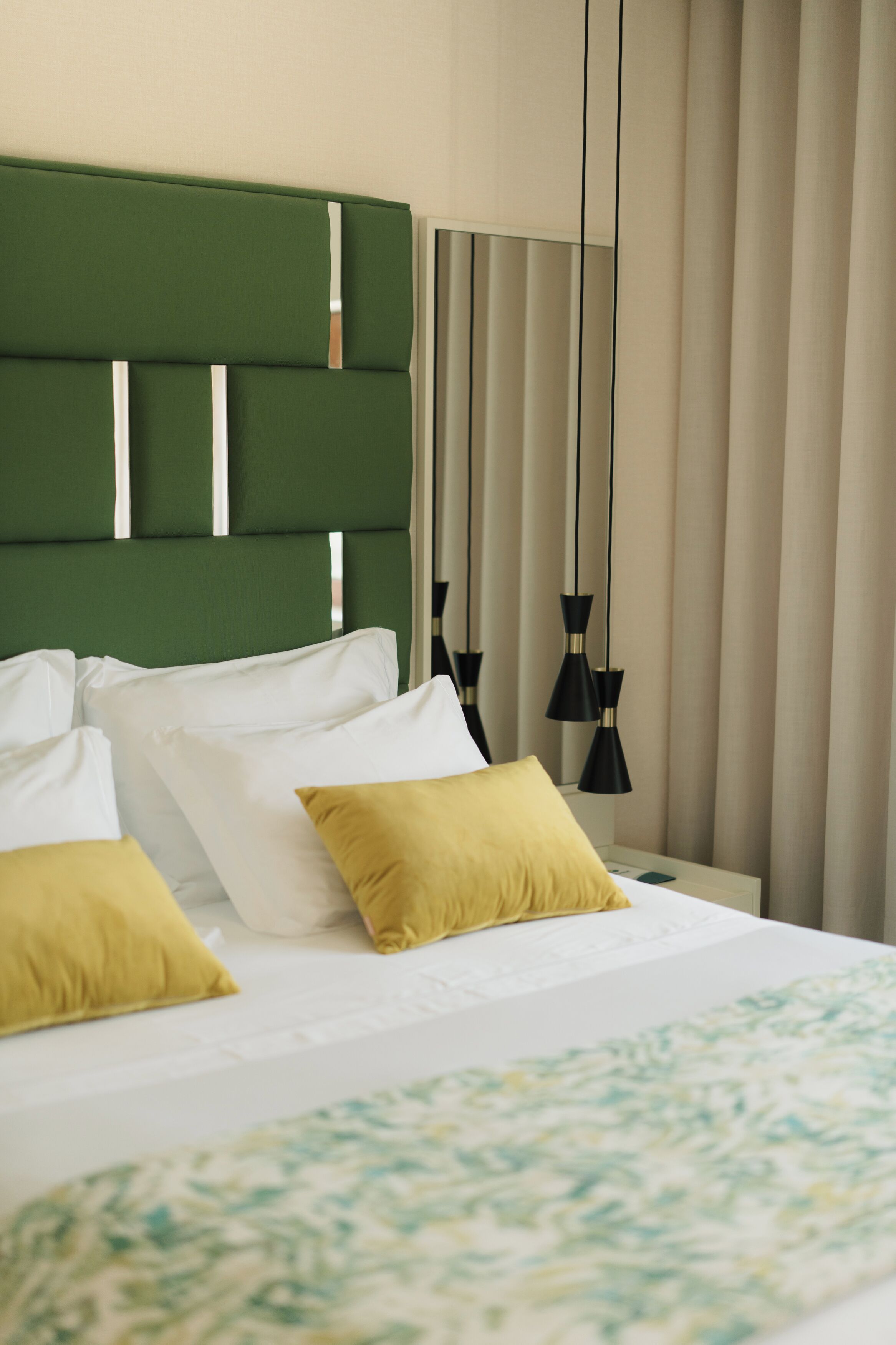 A hotel room with a green headboard, white and yellow pillows, a bed with a patterned bedspread, and black pendant lights.