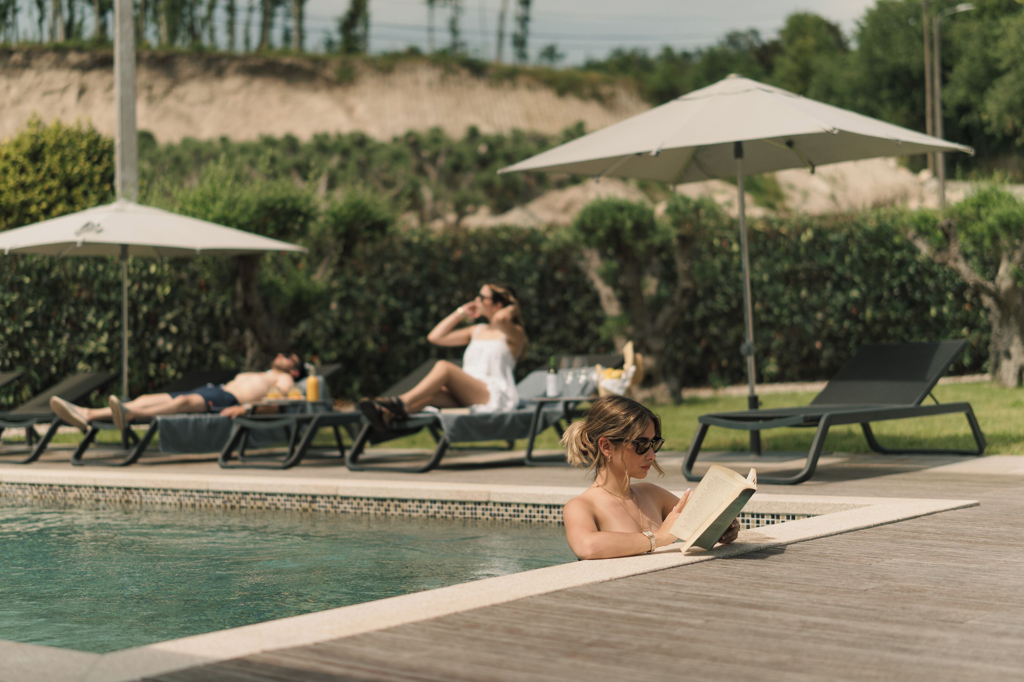 Un área junto a la piscina con sillones de descanso, sombrillas y una persona leyendo un libro.