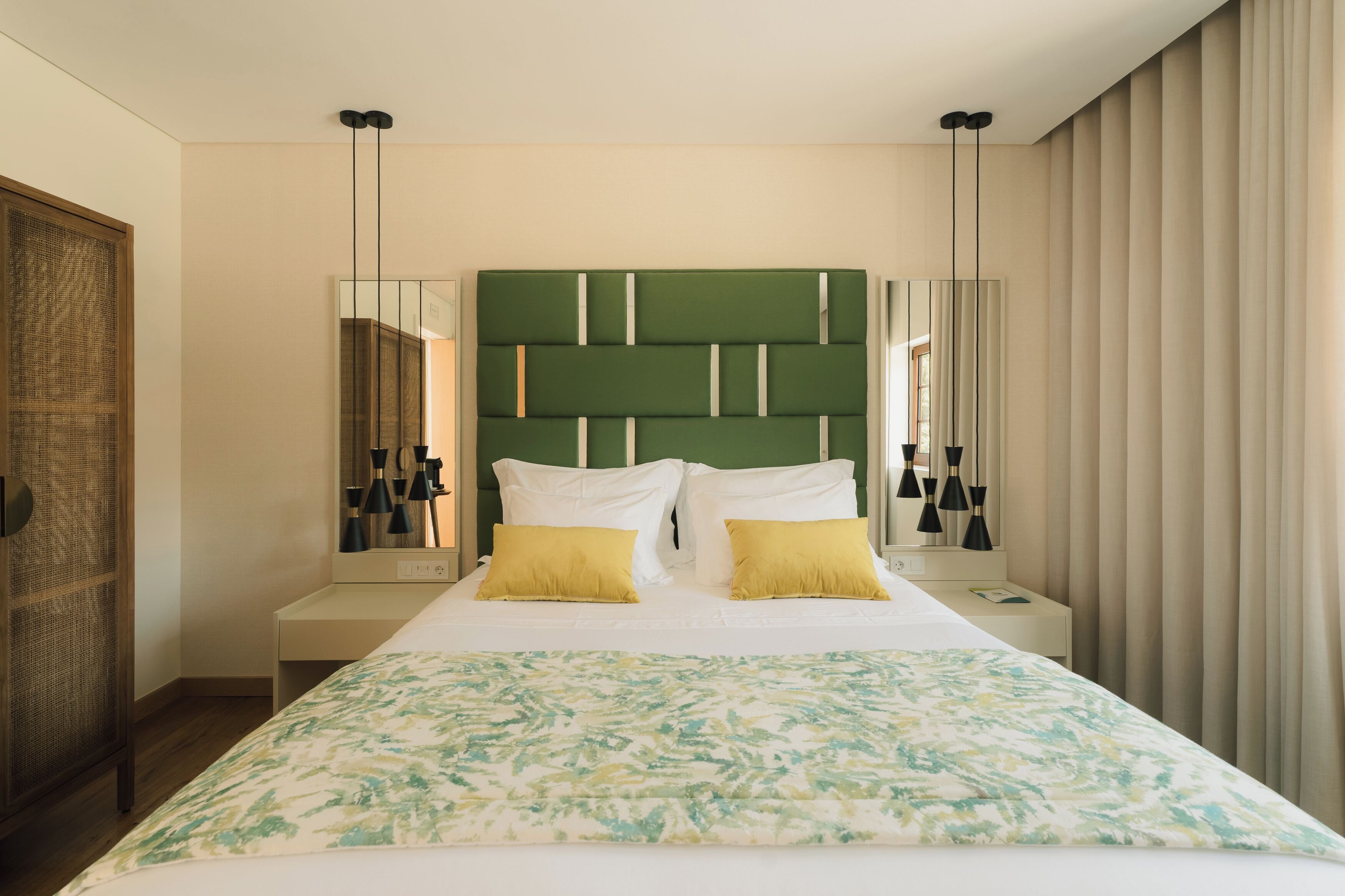 Habitación de hotel con una cama grande, cabecera verde, dos mesitas de noche y un ventanal con cortinas.
