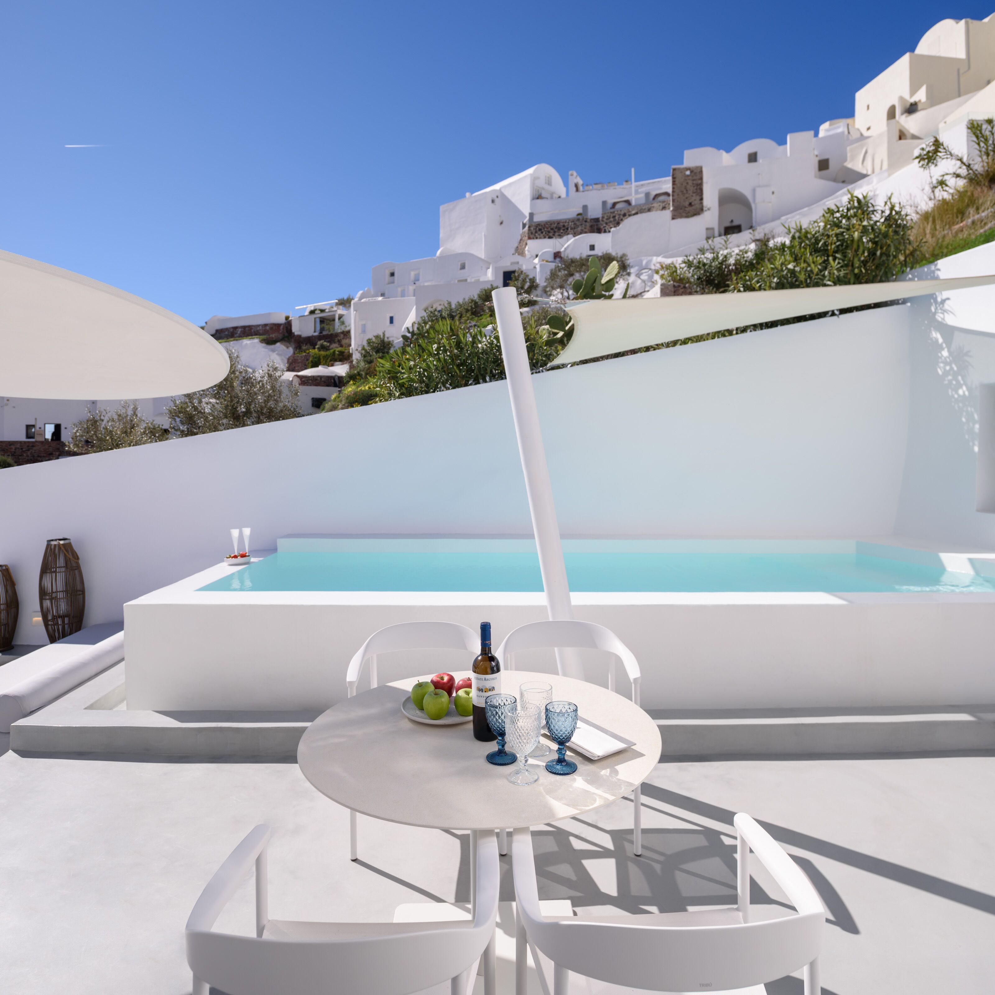 A white outdoor seating area with a table and chairs, a pool, and white buildings in the background.