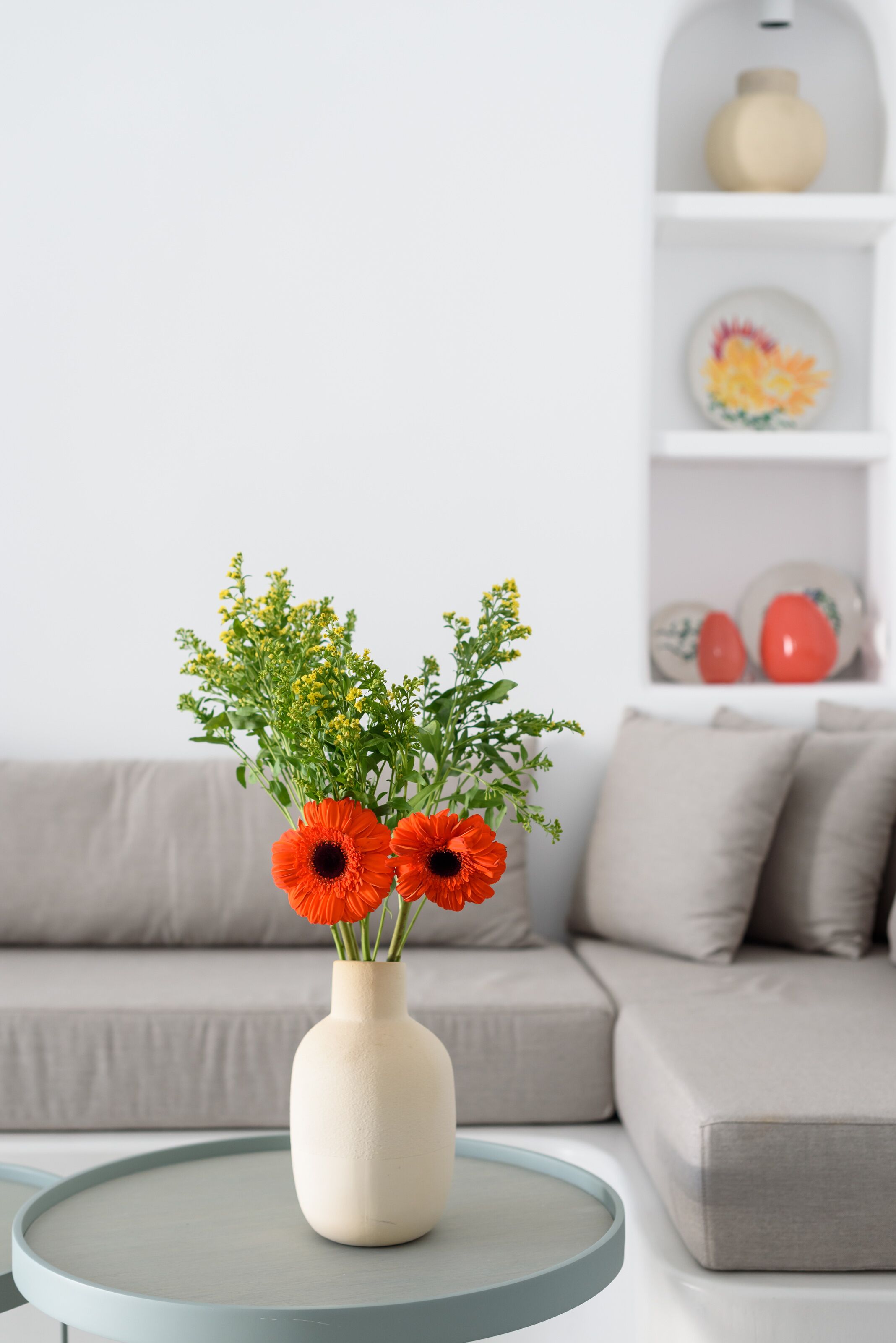 Un salon avec une table ronde, un canapé beige et un vase blanc orné de fleurs.