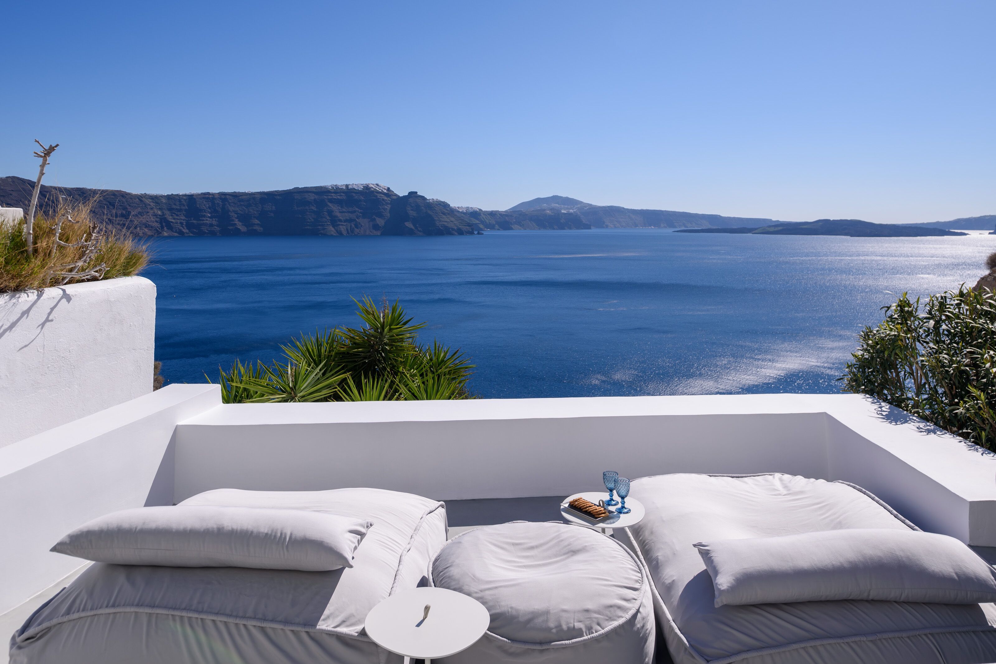 A white infinity pool with two white lounge chairs and a small table, overlooking a clear blue sea and distant mountains.