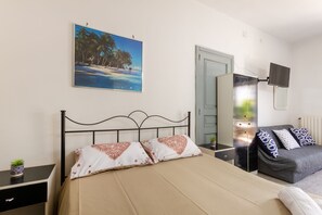 A bedroom with a bed, a sofa, a TV, a painting, and a small plant.