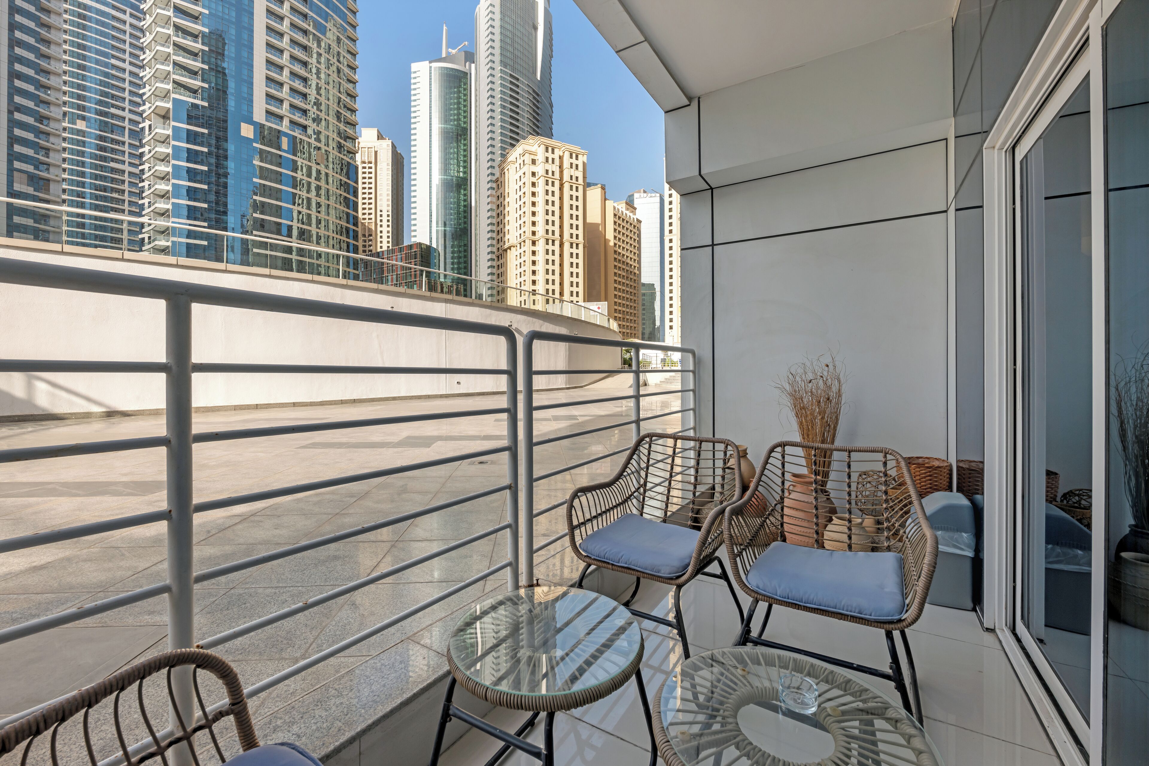 Een balkon met rieten stoelen en een glazen tafel, met uitzicht op een stadsgezicht met hoge gebouwen.