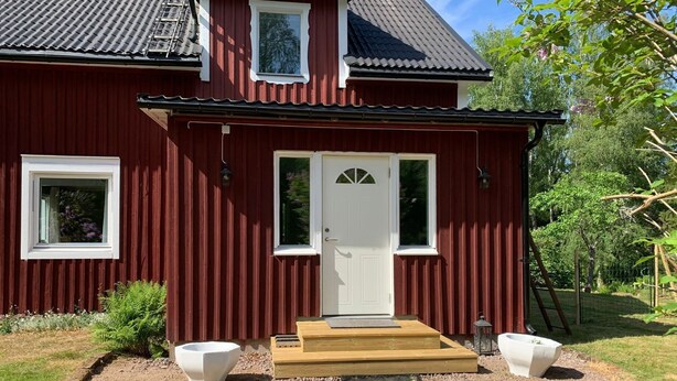 Une maison en bois rouge avec une porte blanche, deux fenêtres et un petit porche avec des marches.