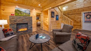 A cozy log cabin interior with a fireplace, comfortable seating, and a staircase leading to another level. - 5BR Cabin Near Parkway | Views, Hot Tub, Theater (Pigeon Forge)